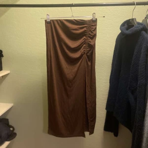 Brun kjol från Gina Tricot - Säljer en snygg brun långkjol från Gina Tricot. Kjolen har en stilren design med en rynkad detalj på sidan som ger en fin effekt. Perfekt för både vardag och fest! 👜 storlek S