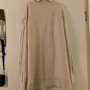 Beige stickad polotröja - Mysig beige stickad polotröja med ett snyggt kabelstickat mönster på baksidan. Perfekt för kyliga dagar med sin långa passform och ribbade kanter. Den har långa ärmar och en hög krage som håller dig varm och stilren.