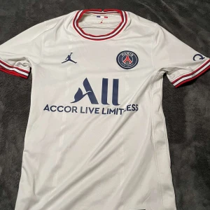 Vit PSG fotbollströja  - Säljer en snygg vit PSG fotbollströja från Jordan. Den har korta ärmar och coola röda och blå detaljer på kragen och ärmsluten. Perfekt för alla PSG-fans som vill visa sitt stöd. Tröjan är i mycket bra skick och redo för nästa matchdag! ⚽