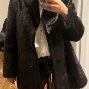 Svart pälsjacka - En lyxig pälskappa köpt i en exklusiv vintagebutik. Pälsen har en naturlig glans och en vacker färg som skiftar mellan mörkbrun och svart, vilket gör den perfekt för att lyfta vilken outfit som helst. Klassisk och tidlös modell som passar både vardag och fest.   Mått: Märkt storlek 48, men uppskattas passa en svensk S eller liten M (ca 36/38). Exakta mått: armhåla till armhåla ca 48 cm och längd ca 77 cm. För bästa passform, jämför gärna måtten med en kappa du redan äger. Nyskick