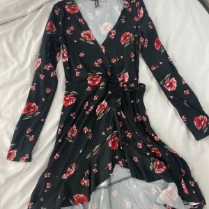 Blommig omlottklänning - Säljer en superfin blommig omlottklänning med v-ringning och långa ärmar. Klänningen har en mörk bas med röda och rosa blommor, perfekt för både vår och höst. Den är i bra skick och passar perfekt för en festlig kväll eller en dag på stan.