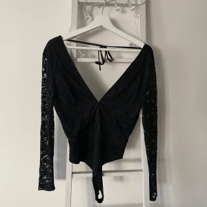 Svart spetsbody från NA-KD - Säljer en  svart spetsbody från NA-KD i storlek 38. Den har långa ärmar och en djup v-ringning med knytband i ryggen. Perfekt för en festlig kväll eller en utekväll med vänner. Den är i mycket bra skick och redo att bäras av någon ny! 🖤