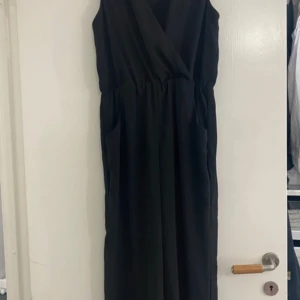 Svart byxdress - Elegant svart byxdress med omlott-design. Perfekt för festliga tillfällen eller en stilren vardagslook. Byxdressen har en v-ringad hals och är ärmlös, vilket ger en sofistikerad touch. Materialet ser ut att vara mjukt och bekvämt, vilket gör den idealisk för både sommar och höst.