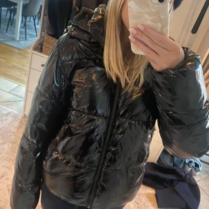 Svart glansig Nelly jacka - Säljer en svart glansig pufferjacka som är perfekt för kalla dagar. Jackan har en dragkedja framtill och en mysig huva. Den är långärmad och är perfekt för vintern och hösten💕