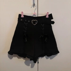 Svarta shorts med hjärtformad detalj från Honey Cinnamon - Säljer ett par supersnygga svarta shorts från Honey Cinnamon. De har en hjärtformat bälte i midjan och snörning framtill. Perfekta för en cool och unik stil! 🖤