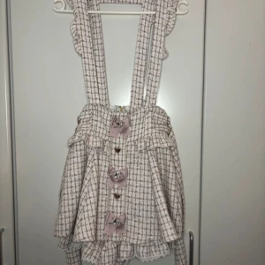 Liz Lisa tweed höngsel kjol  med shorts  - Superfin rutig kjol från Liz Lisa i vitt och rosa (har inbyggda shorts) Den har söta volanger och dekorativa rosetter framtill. Kjolen har   justerbara axelband och en dragkedja samt waist ties bak 💕 strl. Är i deras one size med måtten 60-82 cm i midja och 98 cm i höften!