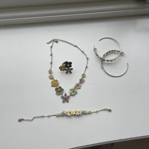 Blommigt smyckeset med halsband, armband och ring - Ett superfint smyckeset med blommor i olika färger. Halsbandet, armbandet och ringen har detaljer i gult, grönt och rosa. Perfekt för att ge en färgglad touch till din outfit. Passar både till vardag och fest!