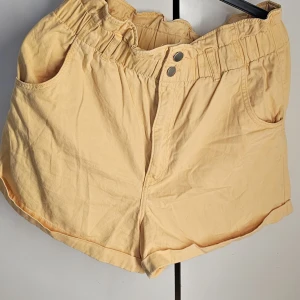 Orange shorts med resårmidja - Säljer ett par bekväma beige shorts i bomull med resårmidja och knappdetalj. Perfekta för sommardagar eller en avslappnad vårlook. De har en lös passform och är i mycket bra skick. Passar perfekt för en dag på stranden eller en chill dag i parken.