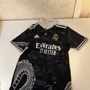 Svart Real Madrid fotbollströja med drakmönster - Säljer en svart Real Madrid fotbollströja från Adidas i mycket bra skick. Tröjan har ett coolt drakmönster i grått och vitt, med klubbens emblem broderat i guld på bröstet. Den är kortärmad med en vit krage och vita detaljer på ärmarna. Perfekt för alla fotbollsfans! ⚽️