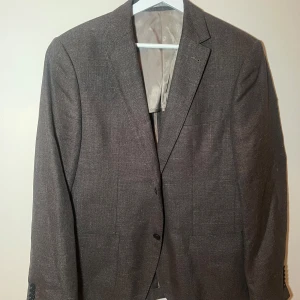 Gråbrun kavaj från Riley - Snygg gråbrun kavaj från Riley i bra skick. Perfekt för både fest och formella tillfällen. Kavajen har en klassisk passform med två knappar framtill och en stilren design. Materialet känns bekvämt och andas bra, vilket gör den idealisk för vår och höst. 