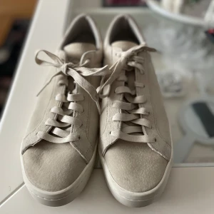 Beige sneakers från Adidas - Säljer ett par stilrena beige sneakers från Adidas i bra skick. De har en klassisk design med snörning och en bekväm sula. Perfekta för vardagsbruk och passar till de flesta outfits. Skorna är använda men i bra skick, en liten fläck framtill på vänster sko samt lite på gummisulan. Herrskor men kan användas av dam. 