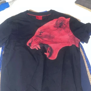 Svart t-shirt med rött lejontryck - Cool svart t-shirt med ett stort rött lejontryck på framsidan. Perfekt för en avslappnad stil eller en utekväll. T-shirten är kortärmad och gjord av mjuk bomull för extra komfort. Passar bra till jeans eller shorts! Pris kan såklart diskuterats 