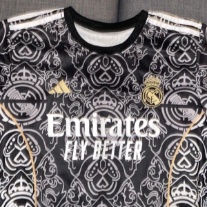 Svart Real Madrid fotbollströja - Säljer en svart Real Madrid fotbollströja med ett unikt mönster i grått och vitt. Tröjan har korta ärmar och är i mycket bra skick. Den har guldiga detaljer och sponsorloggan 'Emirates Fly Better' på framsidan. På baksidan står det 'Mbappé' och nummer 9. 