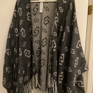 Grå poncho med fransar - Säljer en supermysig grå poncho med ett coolt mönster i ljusgrått. Den har fransar längs nederkanten som ger en bohemisk touch. Perfekt för höst och vår när du vill ha något lätt men ändå varmt. Passar perfekt över en tröja eller blus för extra stilpoäng!