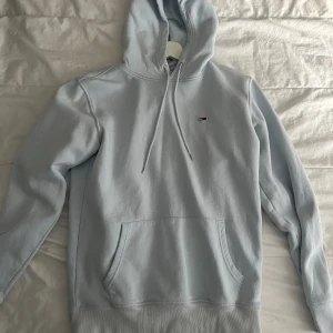Ljusblå hoodie från Tommy Hilfiger - Säljer en stilren ljusblå hoodie från Tommy Hilfiger med en liten logga på bröstet. Den har en klassisk känguruficka och justerbar huva med snören. Perfekt för en avslappnad stil.