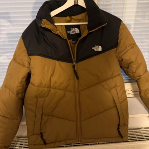 Brun och svart pufferjacka från The North Face - Säljer en snygg brun och svart pufferjacka från The North Face. Jackan har en dragkedja framtill och är perfekt för kyligare väder. Den har långa ärmar och två praktiska fickor med dragkedja. Perfekt för dig som vill hålla stilen i vinter! 🧥