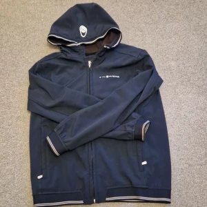 Mörkblå jacka med huva - Snygg mörkblå jacka med huva och dragkedja. Jackan har vita detaljer och ett tryck på bröstet. Perfekt för en casual look.