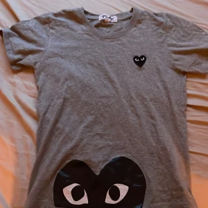 Grå t-shirt från Comme des Garçons - Säljer en grå t-shirt från Comme des Garçons med det ikoniska hjärtmotivet på bröstet. T-shirten har en normal passform och är tillverkad i mjuk bomull. Perfekt för en stilren och avslappnad look. Den passar som xs