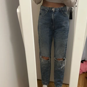 Blå jeans med slitna detaljer - Säljer ett par blå jeans med hög midja och slitna knän. De har en straight passform och klassisk femficksdesign. Perfekta för en avslappnad stil.