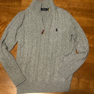 Grå stickad tröja från Polo Ralph Lauren - Säljer en stilren grå stickad tröja från Polo Ralph Lauren med kabelstickat mönster och broderad logga på bröstet. Tröjan har en hög krage och långa ärmar, perfekt för kyligare dagar. Passar både till vardags och mer uppklädda tillfällen. Storlek M även passar som s