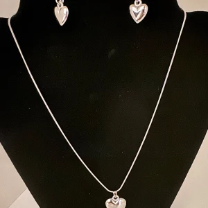 Silverfärgat smyckeset med hjärtan - Charmigt smyckeset i silverfärg med ett halsband och matchande örhängen, båda med hjärtformade hängen. Perfekt för att ge bort på Alla hjärtans dag eller som en kärleksfull present.