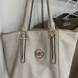 Beige axelväska från Michael Kors - Snygg beige axelväska från Michael Kors med gulddetaljer och kedjehandtag. Väskan har en praktisk dragkedja på baksidan och ett rymligt inre med flera fack och den har en liten fläck på insidan men som går att tvätta. Perfekt för att bära med sig allt man behöver under dagen, till skola eller jobb. 