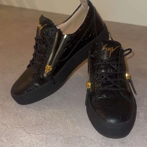 Svarta Guiseppie zanotti öppen för byten - Snygga svarta sneakers med krokodilpräglat mönster och gulddetaljer. Skorna har både snörning och dragkedja för en unik look. Perfekta för att ge din outfit en lyxig touch. Storleken är 40 i italiensk storlek vilket gör att båda du som har på dig 40-41 ska dessa passa dig 
