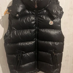 Svart dunväst från Moncler - Snygg svart dunväst från Moncler med dragkedja och huva. Västen har en quiltad design och två fickor med dragkedja framtill. Perfekt för kyligare dagar. MER BILDR KOM PRIVAT! DEN ÄR ÄKTA!