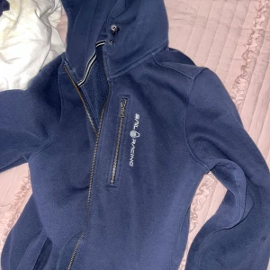 Mörkblå hoodie från Sail Racing - Säljer en mörkblå hoodie från Sail Racing med dragkedja och huva. Jackan har en broderad logga på bröstet och en liten logga på huvan. Perfekt för en casual look.