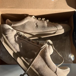 Beige sneakers Polo Ralph Lauren - Snygga beige sneakers från Polo Ralph Lauren i mocka. Skorna har en klassisk design med snörning och en diskret logga på sidan. Perfekta för en stilren look.