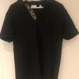 Pike Burberry - Snygg svart Piké från Burberry med klassiskt rutigt mönster på insidan av kragen. Perfekt för en stilren look. Kortärmad och i en bekväm passform.