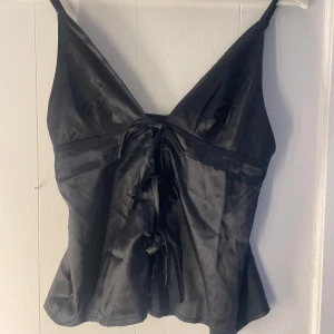 Svart satinlinne med knytning - Snygg svart satinlinne med smala axelband och rosett snörning framtill. Perfekt för en stilren och elegant look. Materialet ger en lyxig känsla och passar bra till både jeans och kjol. Aldrig använd💞💞