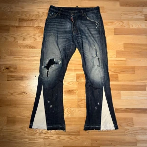DSQUARAD2 ”FLARE JEANS” - Unika blå jeans med slitningar och fransiga kanter. De har en cool slitsdetalj i vitt tyg på benen som ger en edgy look. Perfekta för dig som vill sticka ut!