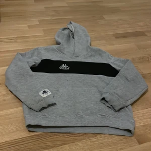 Grå hoodie från Kappa - Säljer en grå hoodie från Kappa med svart band och logga på bröstet. Tröjan har långa ärmar och en klassisk huva. Perfekt för en avslappnad stil.