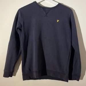 Mörkblå tröja från Lyle & Scott - Säljer en stilren mörkblå tröja från Lyle & Scott Junior. Tröjan har en liten gul logga på bröstet och rund halsringning. Perfekt för en avslappnad look.
