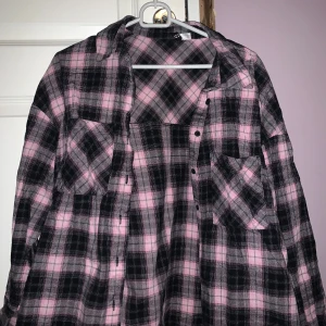 Rutig flanellskjorta i rosa och svart - Säljer en snygg rutig flanellskjorta i rosa och svart. Skjortan har långa ärmar och knappar framtill. Perfekt för en avslappnad stil med en touch av färg.
