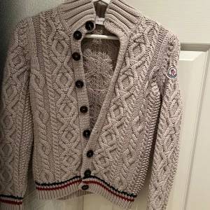 Beige stickad kofta från Moncler - Säljer en snygg beige stickad kofta från Moncler med flätmönster och knappar framtill. Koftan har en hög krage och Moncler-logga på ärmen. Perfekt för kyliga dagar! 🧥