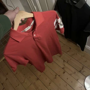 Röd pikétröja från Moncler - ”Snygg röd pikétröja från Moncler med vita detaljer på ärmarna. Perfekt för en sportig look. Tröjan har en klassisk krage och knappar framtill.”🗿 Storlek M 