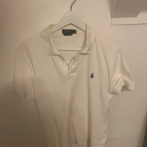 Vit kortärmad pikétröja från Polo Ralph Lauren - Säljer en klassisk vit pikétröja från Polo Ralph Lauren med korta ärmar och en broderad logotyp på bröstet. Perfekt för en stilren och avslappnad look. Tröjan har en krage med knappar.  Äkta