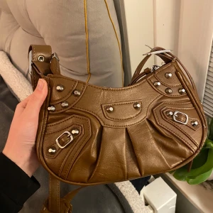 Brun axelväska med nitar - Snygg brun axelväska  med coola nitar och spännen. Väskan har en justerbar axelrem och dragkedja upptill. Även med  innerfickor. Det finns små defekter men inge som är på en gång märkbart eller som hindrar funktionen (fråga för bilder)💞