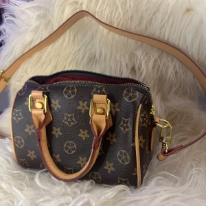 Brun mönstrad axelväska - Snygg brun axelväska med klassiskt mönster och detaljer i beige. Väskan har en dragkedja upptill och ett rymligt rött innerfoder. Perfekt för att bära dina dagliga nödvändigheter med stil.