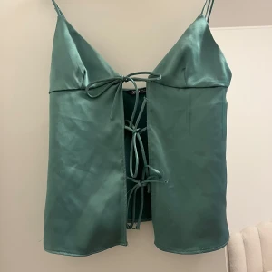 Grön satinlinne från Zara - Säljer denna Snygga gröna tröjan från Zara med smala axelband och knytning framtill. 