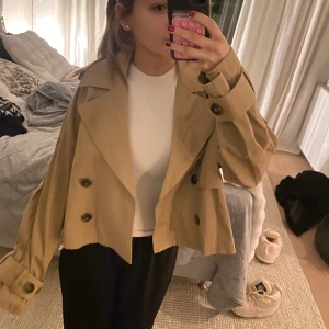 Beige jacka  - Snygg beige trenchcoat från Gina med knappar och klassisk krage. Jackan är croppad och har långa ärmar med dekorativa knappar vid ärmsluten. Helt oanvänd med ett värde på 599kr