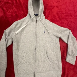 Grå hoodie från Ralph Lauren - Snygg grå hoodie från Ralph Lauren med dragkedja och det klassiska logotypbroderiet i blått på bröstet. Tröjan har en justerbar huva med vita snören och praktiska fickor framtill. Perfekt för en avslappnad stil. Ny pris 2300 kr fler bilder finns vid intresse.