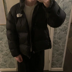 Svart dunjacka från The North Face - Säljer en svart pufferjacka från The North Face. Jackan har en klassisk design med dragkedja och är perfekt för kyligare väder. Den är långärmad och har en hög krage för extra värme. Perfekt för vintern!