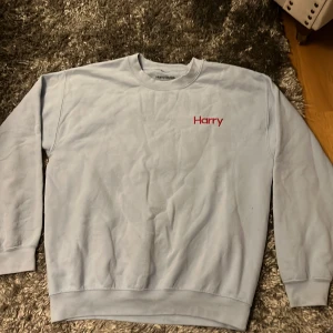 Harry Styles  - Säljer en ljusblå sweatshirt från Harry Styles med röd broderad text 'Harry' på bröstet och 'Treat People With Kindness' på ärmen. Tröjan är tillverkad i en bomulls- och polyesterblandning och har långa ärmar. Rätt bra skicka inga skador/fläckar! Superbra skön tröja och passar till allt🙌🏼 I storlek ”M”