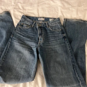 Blå jeans från Bershka - Säljer ett par klassiska blå jeans från Bershka. De har en högmidjad straight passform och är tillverkade i denim. Perfekta för en avslappnad stil. Jeansen har en traditionell femficksdesign och en knappgylf. Slitage vid byxkanten, annars inget mer. 
