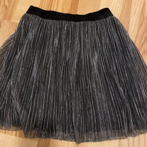 Svart glittrig kjol från Lindex - Snygg svart glittrig kjol från Lindex i storlek 134/140. Kjolen har ett plisserat mönster som ger en fin struktur och en elastisk midja för bekväm passform. Perfekt för att lägga till lite glitter i garderoben!