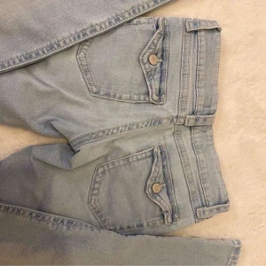 Ljusblå jeans från Gina Tricot - Säljer ett par ljusblå jeans från Gina Tricot med slitna detaljer och dragkedja. De har en klassisk femficksdesign och knappstängning. Perfekta för en avslappnad stil. Strl 152