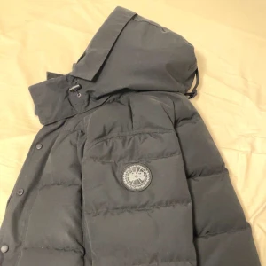 Svart canada goose wyndham black label - Säljer en svart dunjacka från Canada Goose med avtagbar pälsdetalj på huvan. Jackan har en quiltad design och stängs med dragkedja och knappar. Perfekt för kalla vinterdagar. Dock har dragkedjan ett litet problem, inte själva dragkedjan utan saken man tar tag i för att dra ner den. Skriv för mer bilder, köpt för 18 tusen kr, säljs eftersom den ej passar längre och har en ny jacka så denna har ingen betydelse för mig, online kvitto finns.
